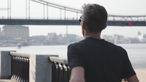 Retrato de homem esportivo em activewear preto correndo no passeio pela cidade. Vista de perto do atleta masculino correndo perto do rio com ponte. Jovem desportivo corre no aterro da cidade. Visão lateral traseira — Vídeo de Stock