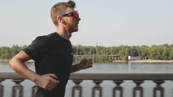 Hombre joven fuerte en camiseta negra y gafas de sol deportivas corriendo a lo largo del río. Un chico activo y enérgico corriendo. Hombre deportivo entrenando en el terraplén de la ciudad. Hombre caucásico haciendo ejercicios al aire libre en muelle de la ciudad — Vídeo de stock