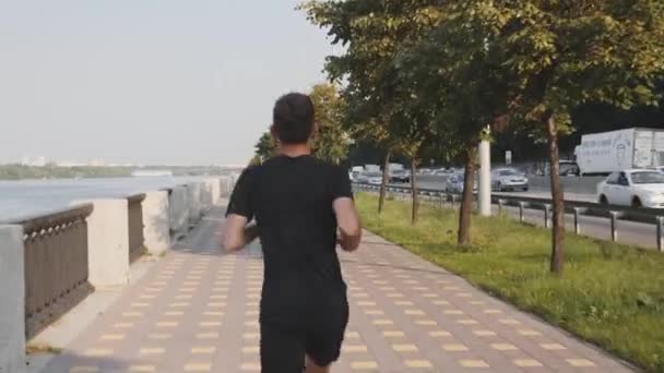Um jovem atlético de t-shirt preta e calções desportivos corre ao longo de um passeio abandonado de manhã cedo. Um tipo activo desportivo a correr num passeio pedestre perto da auto-estrada barulhenta. Slim menino correndo devagar — Vídeo de Stock