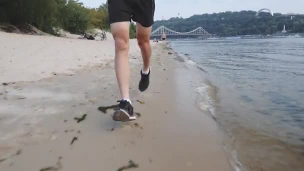 Entrenado musculoso piernas masculinas corre en la playa del río. Vista frontal del hombre deportivo activo corriendo cerca del río. Potentes piernas fuertes corriendo a lo largo del río. Joven caucásico chico haciendo entrenamiento al aire libre . — Vídeos de Stock