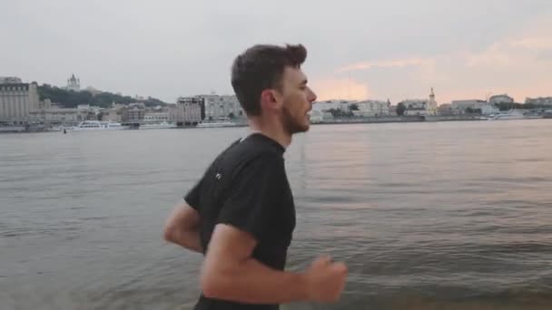 Joven chico caucásico en camiseta negra corriendo en la playa del río. Hombre delgado activo en traje deportivo corre a lo largo del río. El tipo corre cerca del río. Vista lateral del joven corriendo a lo largo del río con la ciudad en el fondo — Vídeo de stock