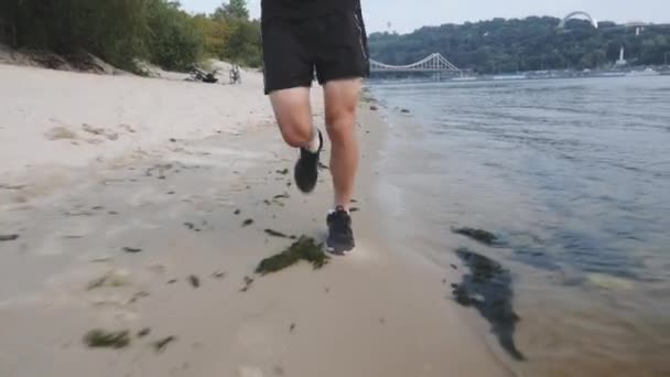 Gambe maschili muscolose allenate corrono sulla spiaggia del fiume. Un giovane concentrato che corre vicino al fiume. Potenti gambe forti che corrono lungo il fiume. Giovane ragazzo motivato che fa allenamento all'aperto. Rallentatore — Video Stock