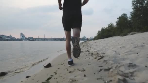 筋肉の強力な男性の足は、川のビーチに沿って実行されます。夕方に川の近くでジョギングするスポーティな男性の足。川の近くでやる気の少年の訓練。小川に沿って走る強い男性の足のクローズアップ。スローモーション — ストック動画