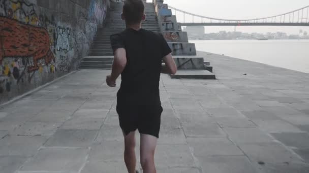 Widok z tyłu profesjonalnego sportowca działającego na nabrzeżu. Sportive Slim człowiek biegnie wzdłuż rzeki. Athletic Fit młody chłopak działa na promenadzie w pobliżu rzeki. Młody facet jogging po schodach. Zwolnionym — Wideo stockowe