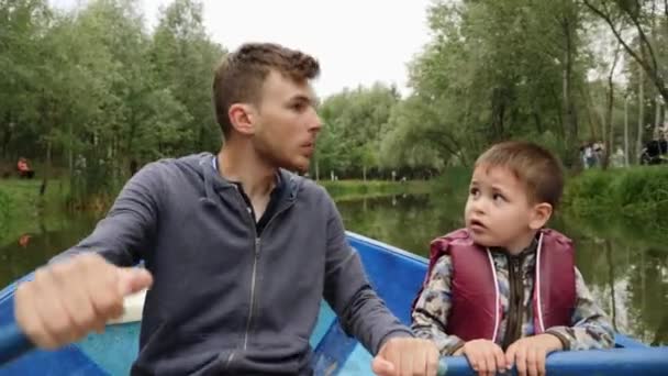 Giovane padre felice vela barca di legno e suo figlio lo aiuta. Padre e figlio remano in barca e parlano tra loro nel parco verde sul lago. Buon papà divertirsi con il suo piccolo figlio carino fuori — Video Stock