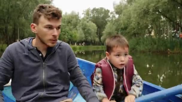 Šťastný otec a syn plaví na jezeře na lodi mezi zelenými stromy. Malé milé dítě, které mluví se svým mladým otcem o jezeře v parku. Malý chlapec, který na lodi veslovat horlivě. Rozkošné dítě s zábavou v parku — Stock video