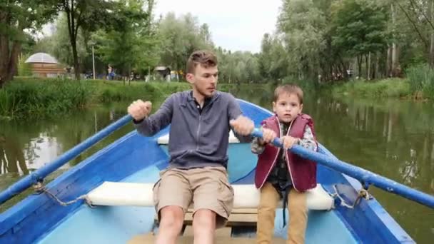 Young father and his little cute son sail on blue boat on lake among green trees. Happy daddy and son spend time together on lake. Happy father and son floating together on blue boat in green park. — Stock Video