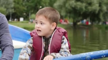 Can yeleği giymiş sevimli küçük çocuk gölde hızlı bir şekilde sıralanmış. Teknede oturan küçük sevimli çocuğun yakından görünümü. Parktaki gölde ahşap teknede kürek çeken komik çocuk. Küçük çocuk göl ile parkta eğleniyor