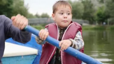Sevimli küçük çocuk konuşuyor ve gölde kamera gülümsüyor. Parkta ahşap teknede yelken sevimli çocuk. Küçük mutlu çocuk kürek yapışıyor ve babası kürek yardımcı olur. Gölde can yeleği komik çocuk