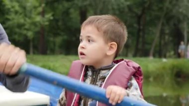 Gölde parkta vakit geçiren küçük sevimli bir çocuğun portresi. Mutlu küçük çocuk teknede oturup etrafa bakıyor. Yakından görüş. Can yeleği sevimli küçük çocuk ahşap tekne oturur.