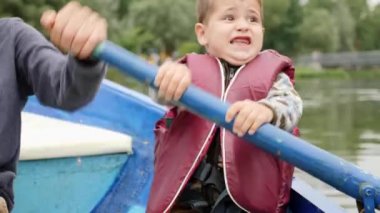 Göldeki parkta özenle kürek çekmek için küçük komik çocuk. Göldeki küçük duygusal çocuğun portresi. Sevimli çocuk teknede oturur ve gülümsüyor. Küçük çocuk yeşil şehir parkında göl üzerinde yüzen.