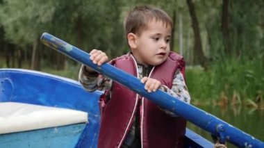 Can yeleği içinde sevimli küçük çocuk teknede oturan ve ahşap kürek tutan. Sevimli çocuk yönlendirmek ve kürek tekne çalışır. Sevimli küçük çocuk parkta teknede yüzen. Gölde teknede çocuk yelken