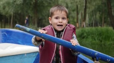 Küçük sevimli çocuk ahşap tekne oturur ve şehir parkında göl üzerinde kürek tutar. Sevimli çocuk gölde tekne kürek çalışır. Parkta teknede yüzen can yeleği küçük çocuğun yakından görünümü.