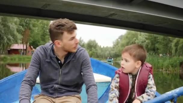 Giovane padre felice e il suo piccolo figlio carino galleggiante su una barca di legno sotto il ponte pedonale e alzando le mani. Famiglia felice divertirsi sul lago. Giovani papà e figlio navigano in barca di legno nel parco — Video Stock