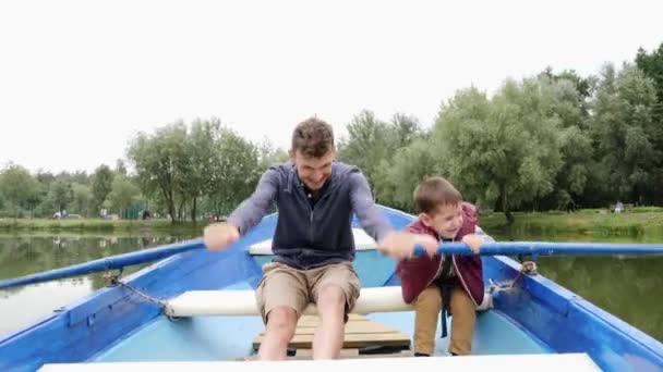 幸せな父と息子は公園の湖でボート旅行を楽しんでいます。幸せな家族は屋外で一緒に時間を過ごします。お父さんと子供がボートで一緒に遊んでいます。笑顔の息子と一緒にボートに浮かぶ陽気な父親 — ストック動画
