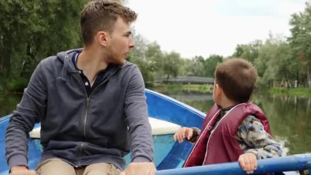 Portret van jonge gelukkige vader en kleine zoon op houten boot in Park. Vader en klein kind rusten in Park op boot en glimlachend. Gelukkige vader en zoon drijven op boot in Park en tijd samen doorbrengen — Stockvideo