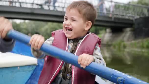 Portret adorable małego dziecka śmiać się i uśmiechać się nad jeziorem. Szczęśliwy mały chłopiec siedzi w łodzi i brodzik. Z bliska widok szczęśliwy uśmiechnięty chłopak na jeziorze w parku. Little Cute dziecko trzyma wiosło — Wideo stockowe