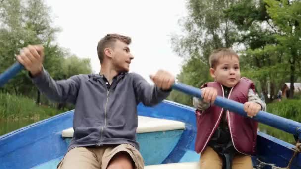 Šťastný otec a syn drží pádla a veslovat na lodi na jezeře. Šťastný otec a malé dítě mluví a usmívají se. Mladý otec a malý syn vznášející se na dřevěné lodi u jezera v zeleném parku. — Stock video