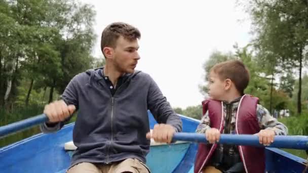 Junger Vater sitzt mit kleinem Sohn auf einem Boot auf dem See. junger glücklicher Vater, der mit seinem kleinen entzückenden Kind auf einem Holzboot im Park schwimmt. Vater und Sohn segeln mit Boot auf See im Park — Stockvideo