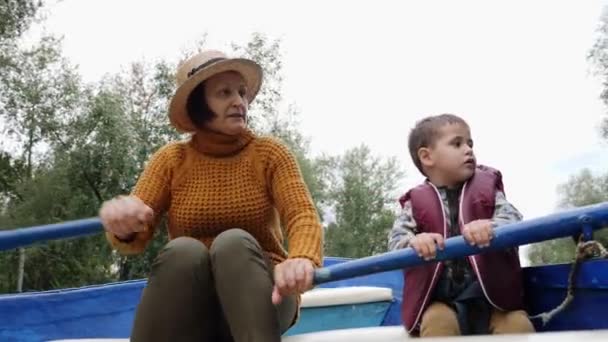 公園の湖に孫と一緒に青い木製のボートに浮かぶ祖母。公園でボートに乗って幸せな家族。おばあちゃんと孫が話し、都市公園の湖の上に木製のボートに座って — ストック動画