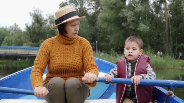 Malé roztomilé dítě v záchranné kazajce plachtí s babičkou na dřevěném člunu na jezeře. Rozkošný chlapec plující s babičkou na lodi. Malé dítě pluje s babičkou na jezeře. Dítě a babička v parku — Stock video