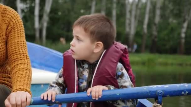木製のボートにパドルを持つ小さな男の子。おかしな子供はボートに座ってオールを保持します。小さな子供がボートに浮かび、周りを見回す。公園の湖に青いボートに浮かぶライフジャケットのかわいい小さな子供 — ストック動画