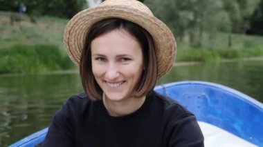 Hasır şapkalı genç esmer kız kameraya gülümsüyor. Gölde ahşap teknede yelken çekici kız. Şehir parkında teknede yüzen kız. Mutlu kız kameraya gülümsüyor. Gülümseyen kız kameraya utangaç