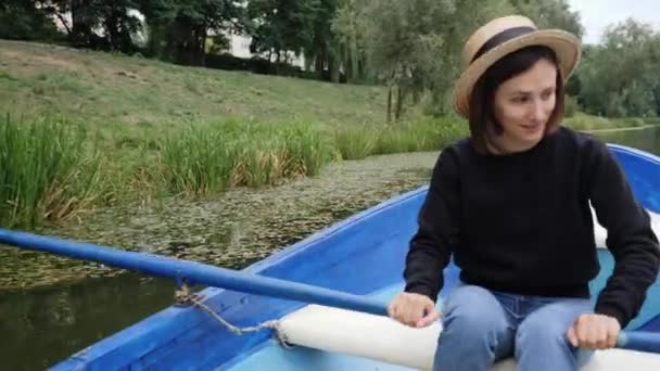 Joven chica feliz en sombrero retro flotando en barco de madera azul en el lago con nenúfares. Chica tímida remando remos en el lago en el parque de la ciudad. Sonriente joven navegando en barco de madera a lo largo del parque de la ciudad — Vídeo de stock