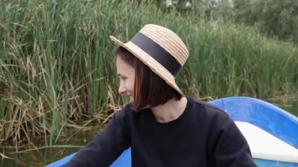 Молодая кавказская девушка в соломенной шляпе гребёт на лодке и улыбается. Счастливая девушка развлекается в парке на озере. Привлекательная брюнетка в соломенной шляпе на деревянной лодке. Девочка-кузнец садится на лодку в парке — стоковое видео
