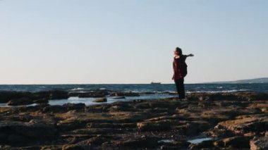 Hipster ekose kırmızı gömlek li genç kadın sahil kenarında duruyor. Rüzgarlı bir günde kayalık sahilde duran elleri ile mutlu genç kadın. Kıyıkenarında duran kıza rüzgar eser