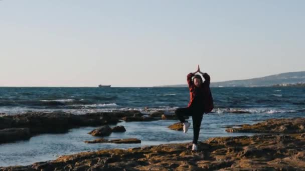 Jovem em roupas casuais está fazendo pose de ioga na praia. Mulher feliz fazendo pose de meditação. Mulher em pé em meditação posar na praia. Mulher sorridente divertida na praia. Menina se divertindo na praia . — Vídeo de Stock