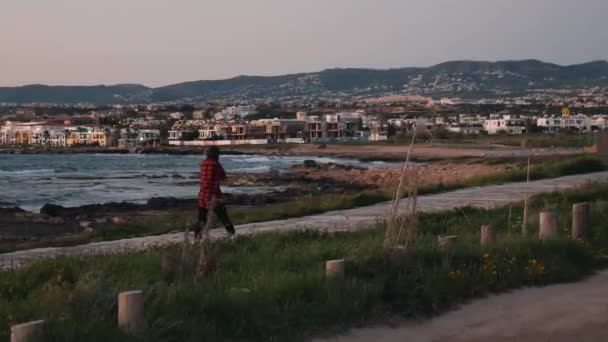 Jonge vrouw in Plaid rood shirt wandelen langs lege kust in de avond. Aantrekkelijke brunette vrouw die op een lege promenade langs de Middellandse Zee loopt. Vrouw lopen op Paphos Quay — Stockvideo