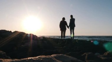 Genç romantik çiftin silueti gün batımında sahilde duruyor. Adam sevgili kadın elini tutar, onunla konuşurken ve denizde smth gösteren. Deniz kıyısında gülen ve gülümseyen genç çiftin silueti