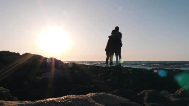 Silueta přítele a přítelkyně stojí na kraji pláže a dívá se na horizont. Mladý šťastný pár stojí na břehu, objímá a dívá se na západ slunce. Romantický večer na pobřeží — Stock video