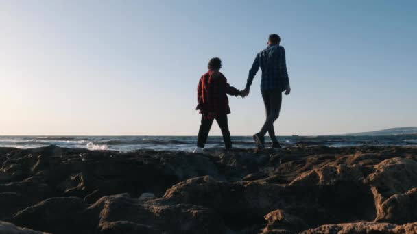 恋爱中的年轻夫妇沿着岩石海岸散步。可爱的家伙和他的女孩站在海边拥抱，看着距离。幸福的夫妇一起散步。男人和女人在海上共度时光 — 图库视频影像