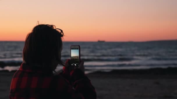 Nahaufnahme Rückansicht einer jungen Frau, die Fotos und Videos von schönen Sonnenuntergängen am Meer macht. attraktive Frau macht Fotos, die bei Sonnenuntergang an der Küste stehen. Frau fotografiert Strand bei Sonnenuntergang — Stockvideo