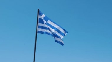 Arka planda mavi berrak gökyüzü ile rüzgarsal sallanan Yunan bayrağı yakından görünümü. Havada Yunan bayrağı dalgalanıyor