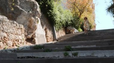 Yaz sarısı elbiseli sevimli kadın beton merdivenlerden aşağı yürüyor, eliyle şapka tutuyor ve yerinde dönüyor