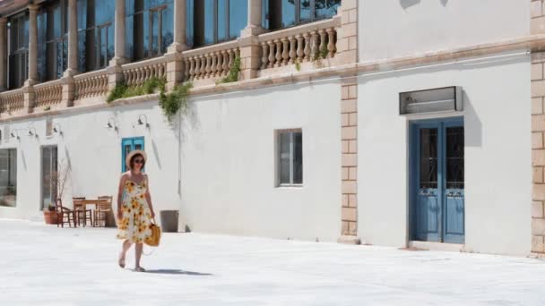 Jonge brunette vrouw in jurk met zonnebloemen en gele handtas wandelen langs lege straat in het historische en Resort deel van de stad — Stockvideo