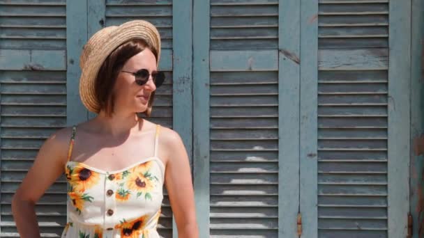 Portret van jonge charmante vrouw in jurk met zonnebloemen en stro hoed staande tegen blauwe venster met jaloezieën en poseren — Stockvideo
