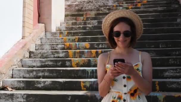Молодая женщина с брекетами в соломенной шляпе и черными солнцезащитными очками стоит на ступеньках с телефоном в руках и улыбается — стоковое видео