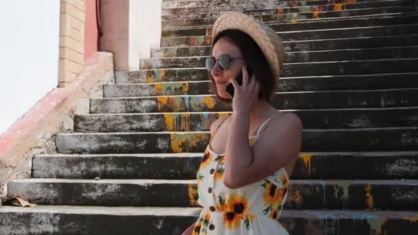 Attraente donna bruna in abito con girasoli in piedi vicino alle scale, parlando al telefono e sorridendo — Video Stock