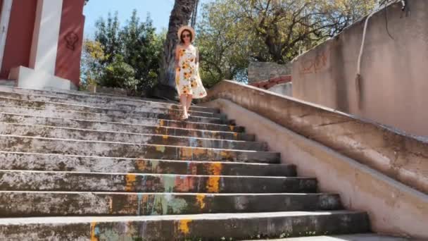 Молодая привлекательная женщина в платье с подсолнухами и соломенной шляпой спускается по лестнице в старом городе. Девушка в отпуске — стоковое видео