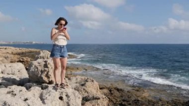 Çekici genç kadın deniz kıyısında kaya üzerinde duran ve telefona bakıyor. Elinde telefon ile deniz kenarında duran Kafkas esmer kız