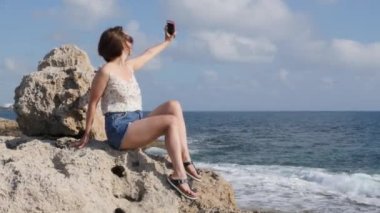 Sıcak esmer kadın pantolon şort kayalık sahil akıllı telefonda selfie yapma. Çekici kadın deniz kıyısında kendini fotoğraf çekmek