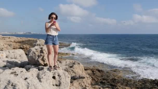 Mujer atractiva caucásica de pie en el borde del acantilado y escribir mensaje en el teléfono. Mujer joven mensajes de texto en el teléfono inteligente en la orilla del mar — Vídeo de stock