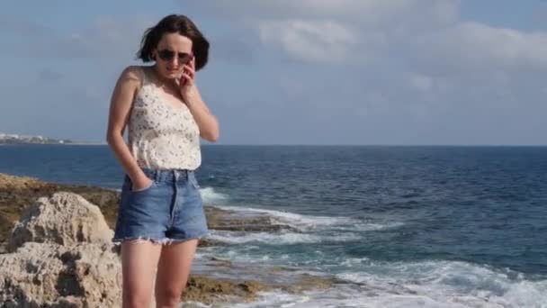 Brunette jonge vrouw in jeans shorts en Silk top staande op de rand van de rotsachtige kust, praten over de telefoon en glimlachend — Stockvideo