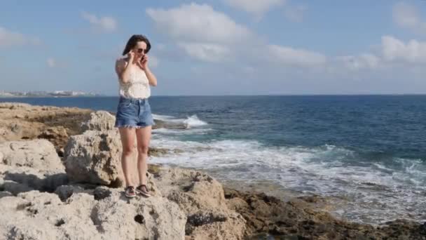 Bonito caucasiano jovem morena em calções jeans e de pé na borda da praia e falando ao telefone — Vídeo de Stock