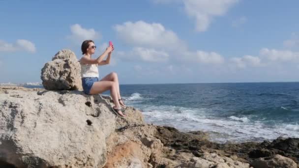 年轻的有吸引力的黑发女人在牛仔裤短裤和丝绸上衣坐在海边的石头上，并拍摄海洋的照片 — 图库视频影像