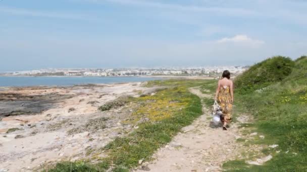 Jeune jolie femme caucasienne en robe d'été avec sac de plage marchant le long du bord de mer. Mignon brunette femelle va sur la plage — Video